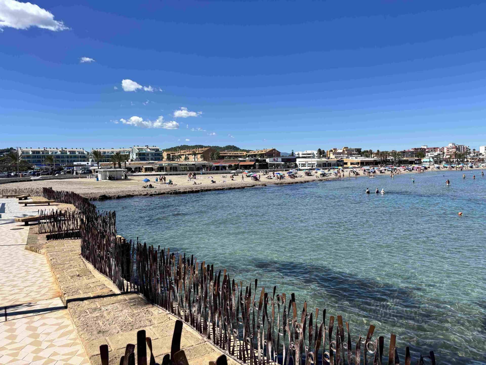 Vibrant Javea beach scene with clear blue waters, sandy shoreline with sunbathers, and lively beachfront restaurants under a bright sky.
