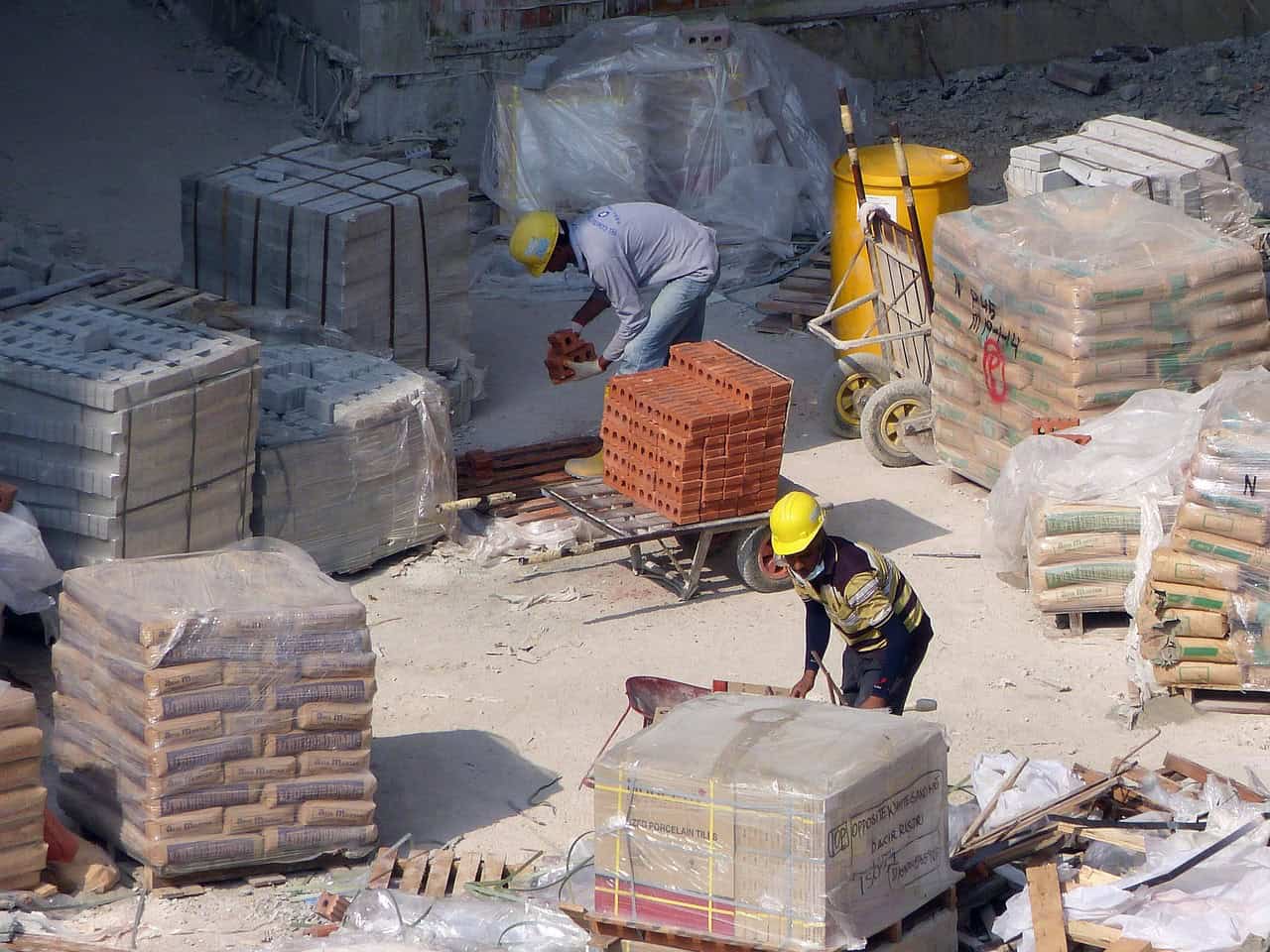 Construction workers on a site with bricks, cement bags, and building tools.