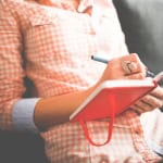 A woman in casual checkered shirt writing in a red notebook with a pen, sitting on a couch at A Place in Javea, offering a relaxing environment for travel inspiration or personal reflection.