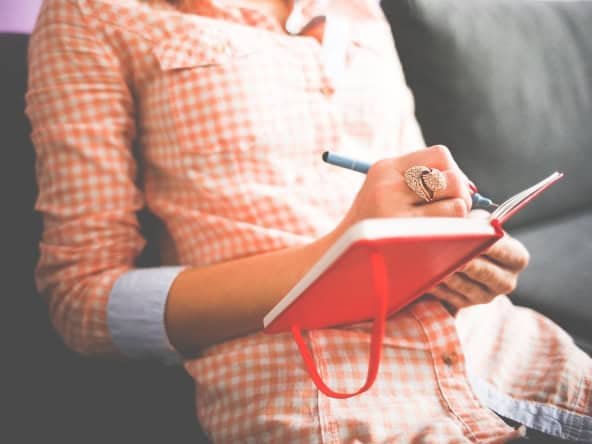 A woman in casual checkered shirt writing in a red notebook with a pen, sitting on a couch at A Place in Javea, offering a relaxing environment for travel inspiration or personal reflection.