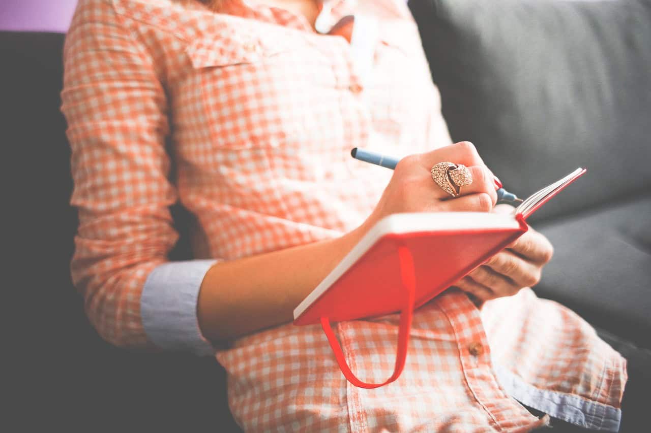 A woman in casual checkered shirt writing in a red notebook with a pen, sitting on a couch at A Place in Javea, offering a relaxing environment for travel inspiration or personal reflection.